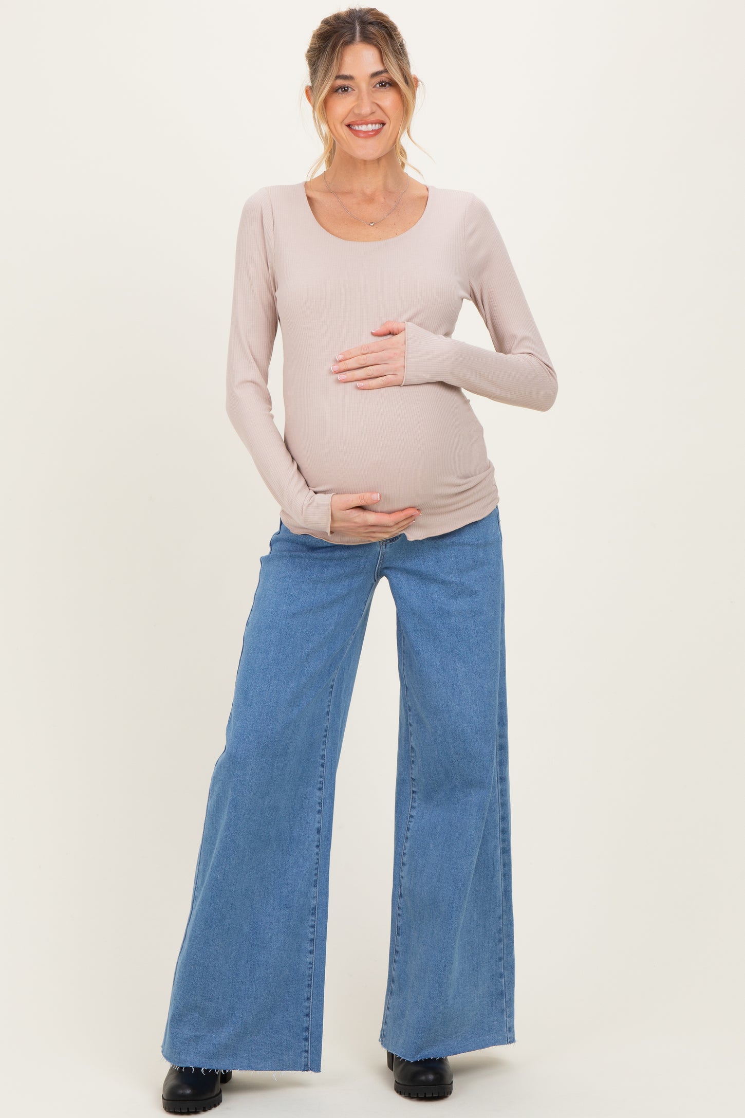 woman wearing a light pink long sleeve maternity top and blue wide leg jeans, holding her baby bump