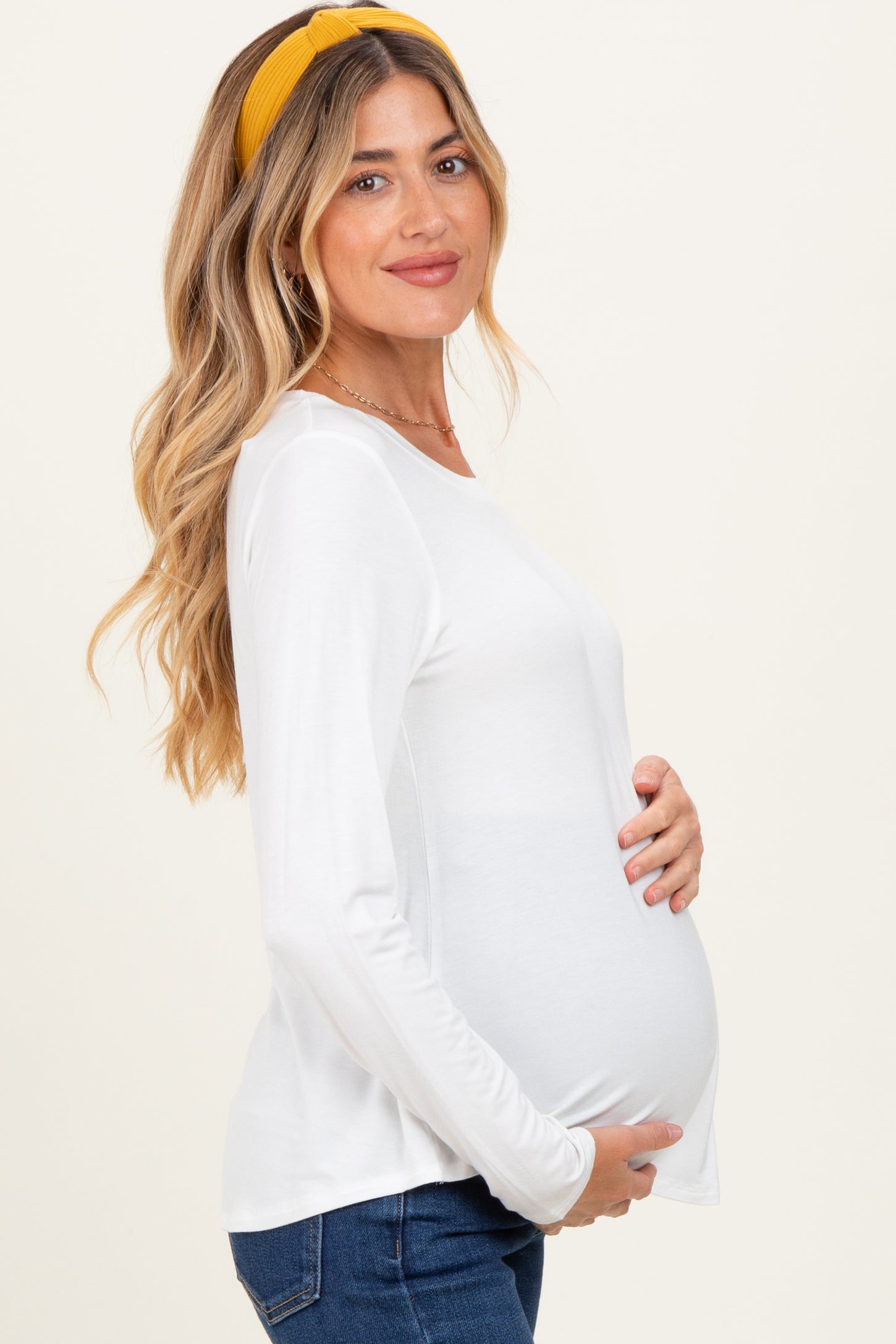 woman wearing a white long sleeve maternity top and holding her baby bump, styled with a yellow headband