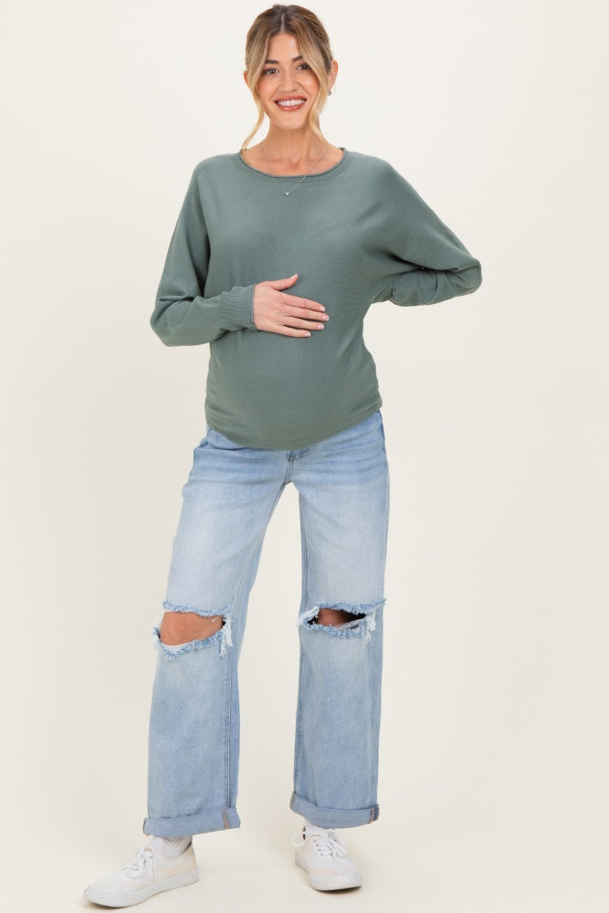 woman wearing a green long sleeve maternity top and distressed light blue jeans, holding her baby bump