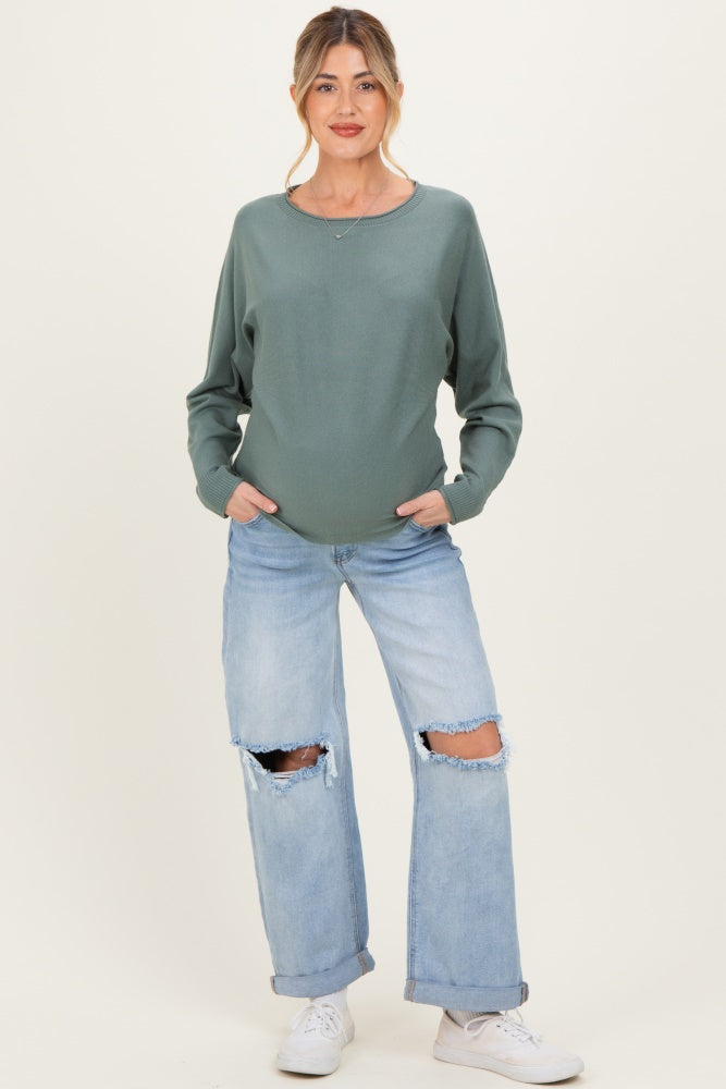 woman wearing a green long sleeve maternity top and distressed light blue jeans, displaying her baby bump