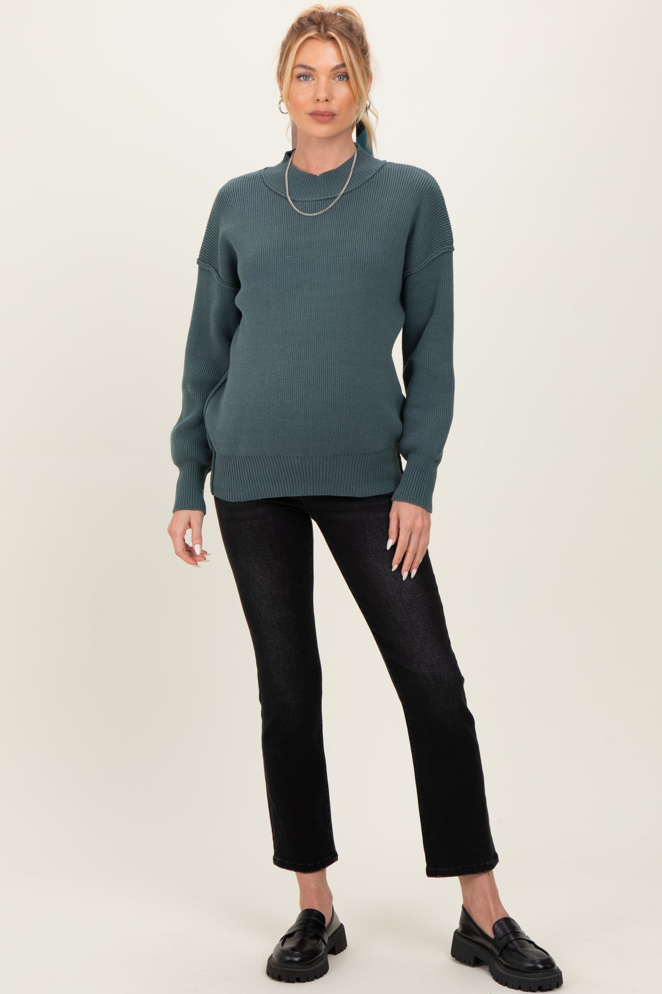 woman wearing a dark teal maternity sweater and black jeans, showing her baby bump, with a layered necklace