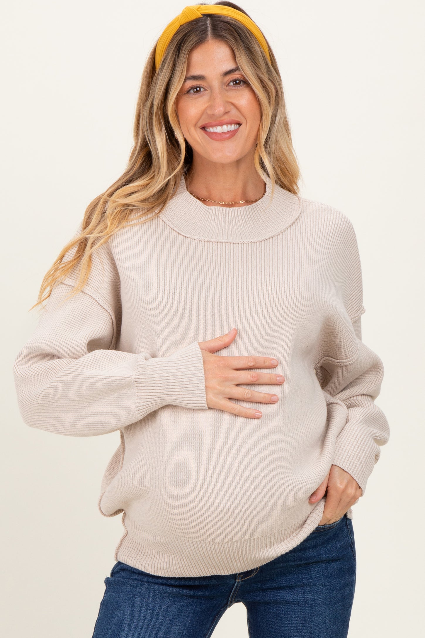 woman wearing a light beige oversized maternity sweater, smiling and holding her baby bump