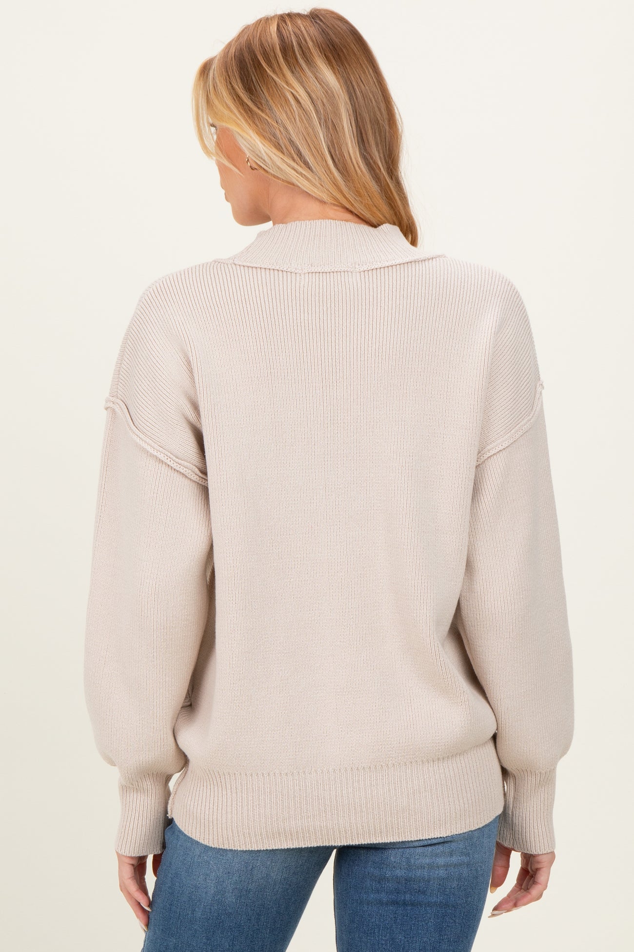 woman wearing a beige oversized knit maternity sweater, view from the back, paired with blue jeans