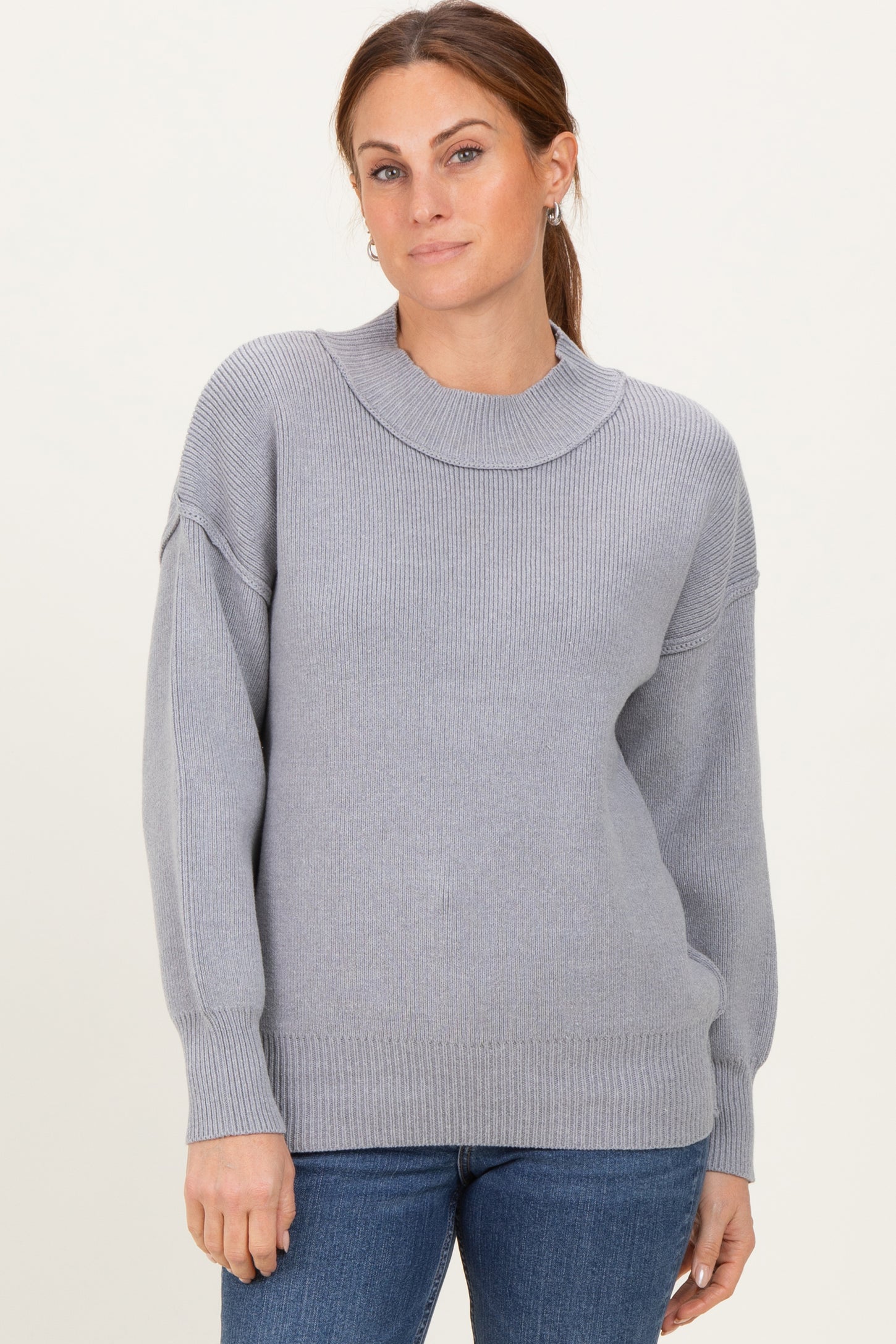 woman wearing a light gray ribbed knit sweater with a high neckline and relaxed fit, paired with blue jeans