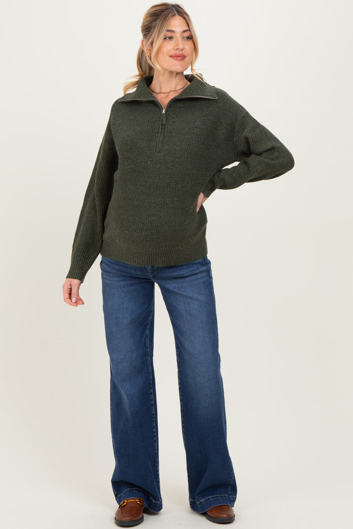 woman wearing a dark green maternity zip-up sweater and flared jeans, posing with a relaxed expression
