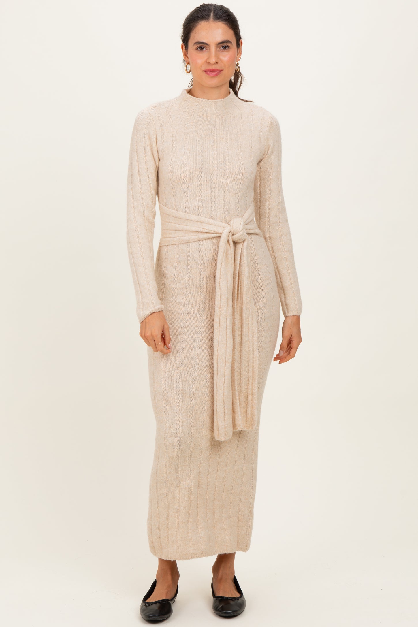 woman wearing a light beige long sleeve knit dress with a tied waist and a straight fit, standing against a neutral background