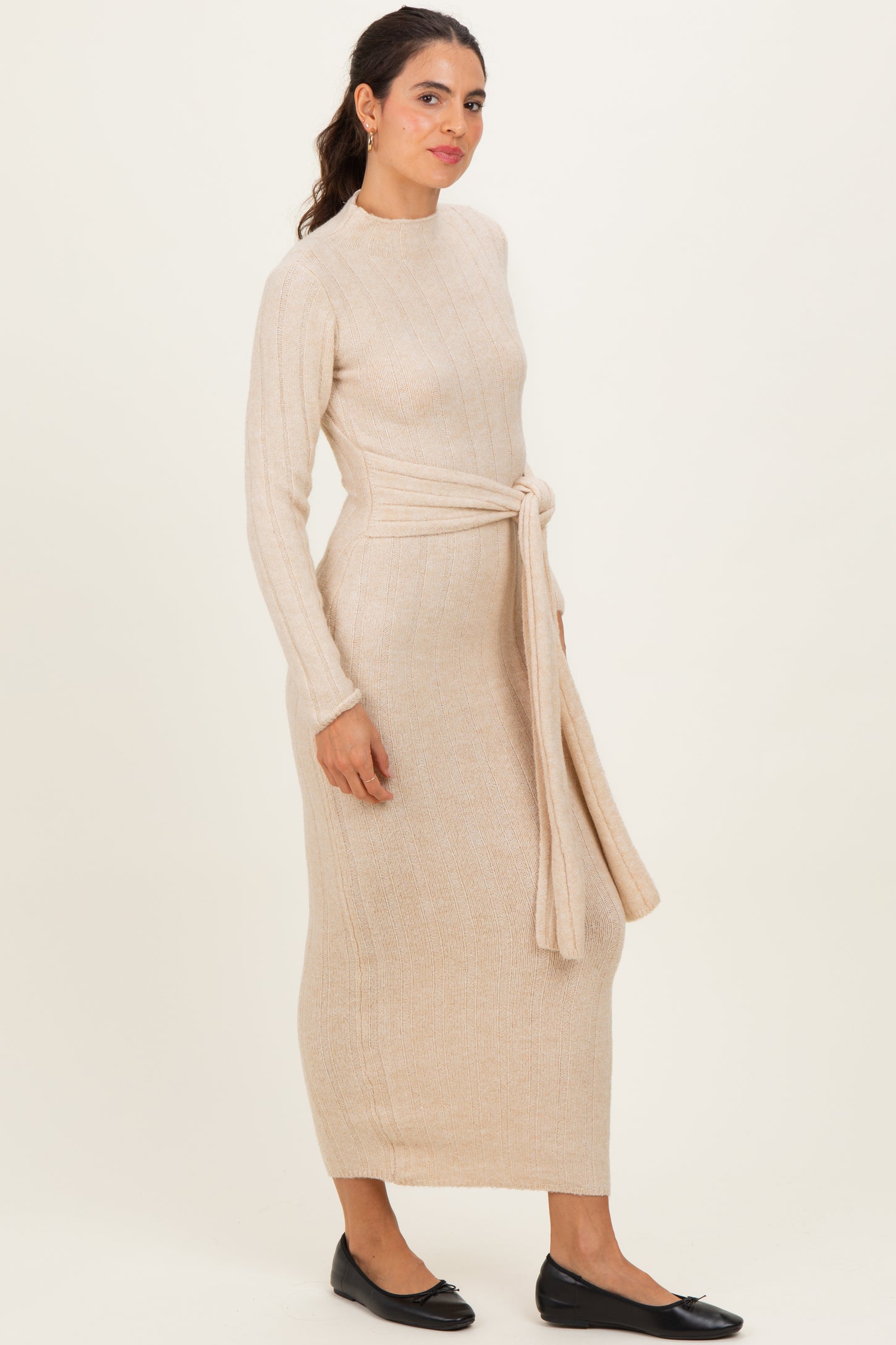 woman wearing a beige ribbed knit maxi dress with long sleeves and a waist tie, standing in profile
