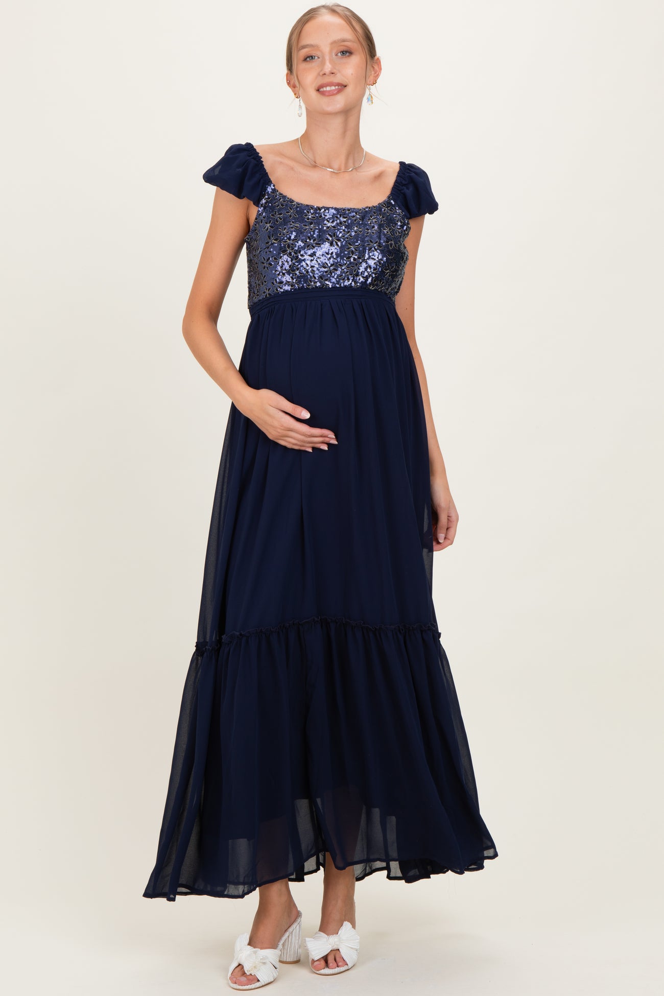 woman wearing a navy blue maternity maxi dress with sequin bodice and ruffled skirt, holding her baby bump