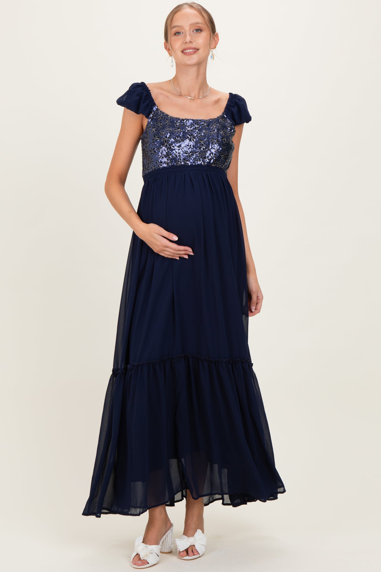 woman wearing a navy blue maternity maxi dress with sequin bodice and ruffled skirt, holding her baby bump
