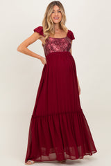 woman wearing a burgundy maternity maxi dress with sequined bodice and ruffled hem, showing her baby bump