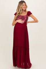 woman wearing a burgundy maternity maxi dress with a sequin bodice and ruffled sleeves, holding her baby bump