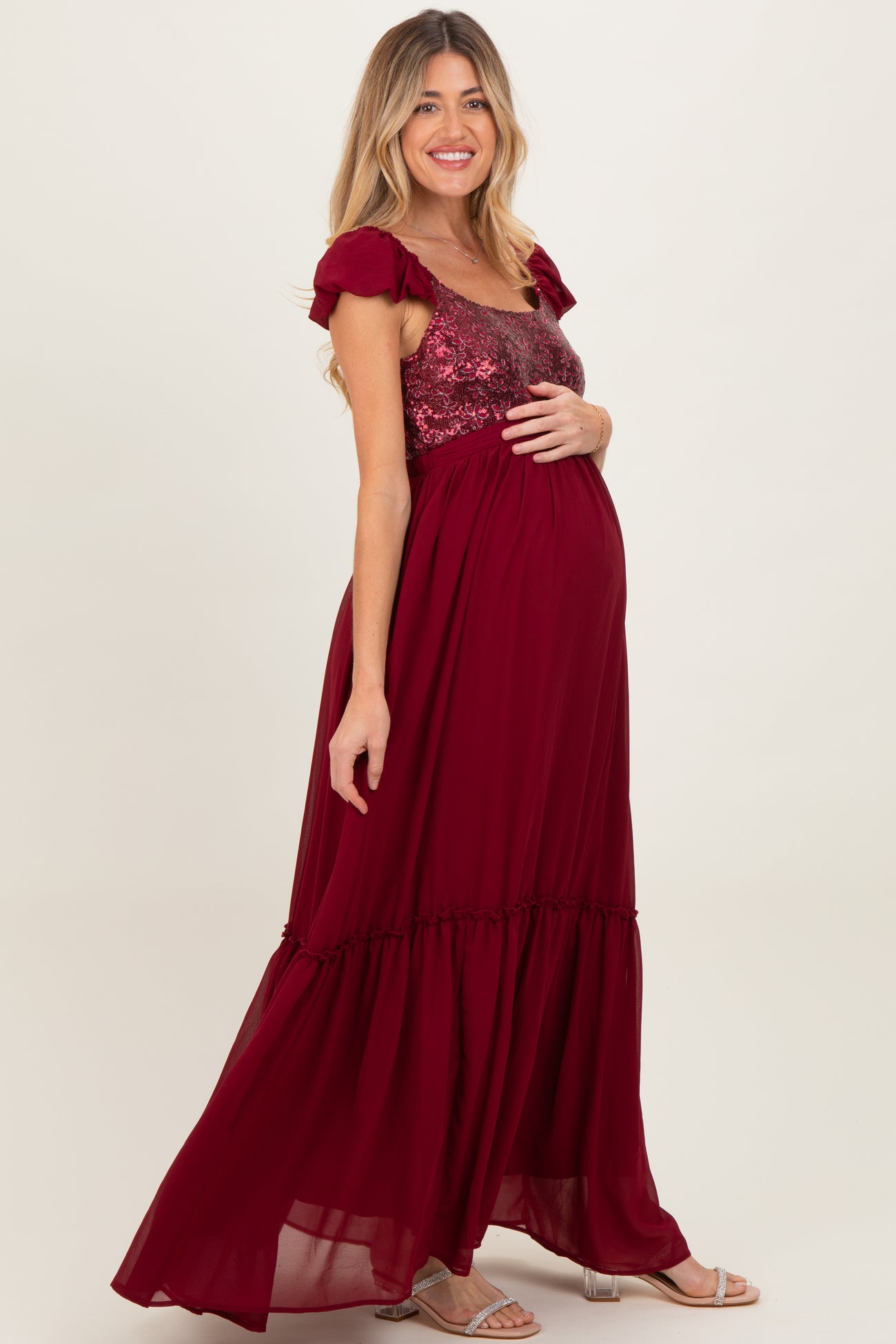 woman wearing a burgundy maternity maxi dress with sequined bodice and ruffled sleeves, gently holding her baby bump