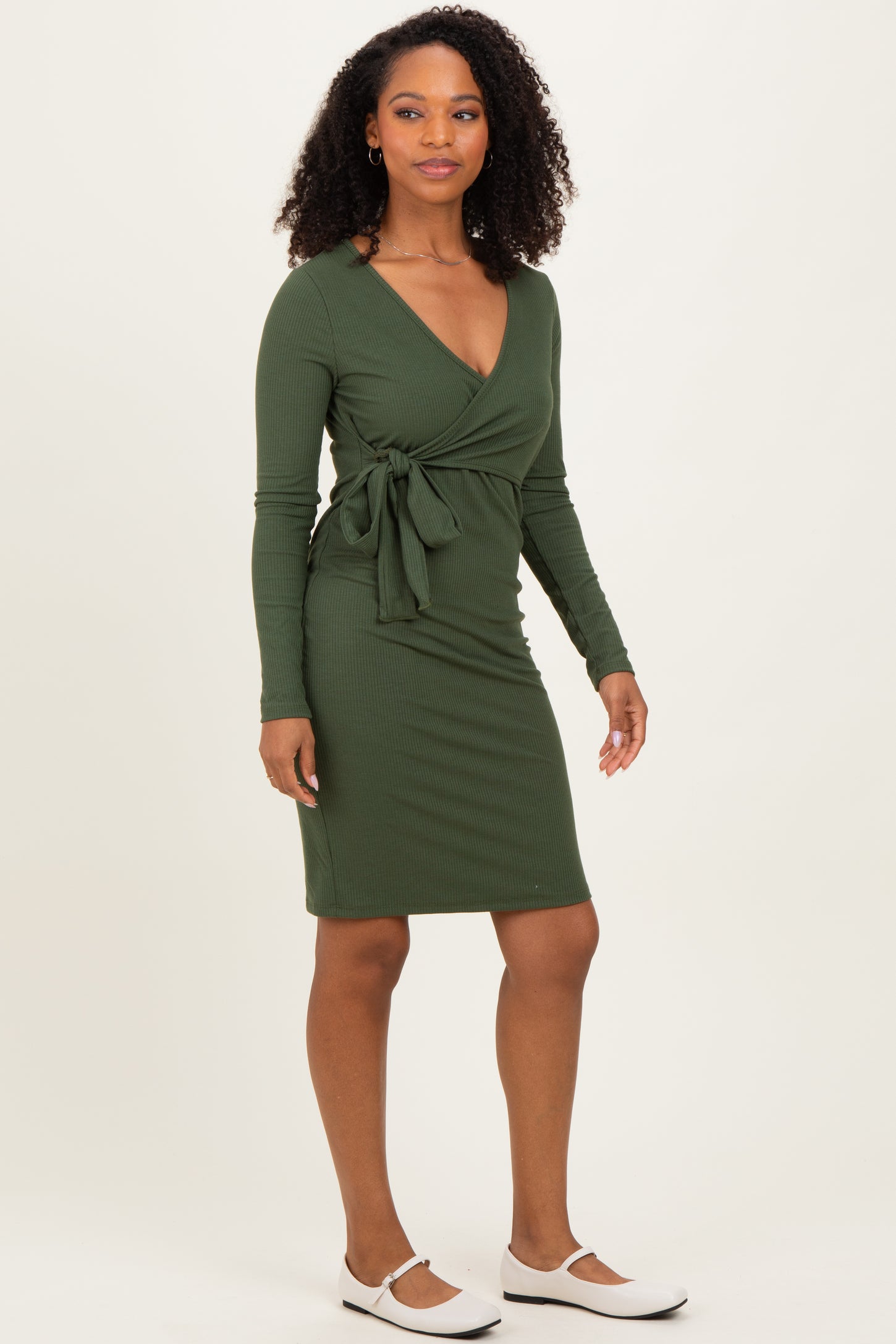 woman wearing a dark green ribbed wrap dress with long sleeves and a tied waist, standing on a light background