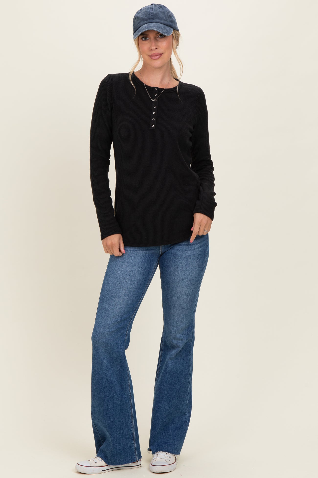 woman wearing a black long sleeve henley top with button details and blue flared jeans, accessorized with a baseball cap