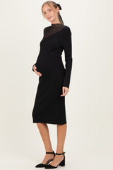 woman wearing a black maternity ribbed dress with a sheer neckline and holding her baby bump