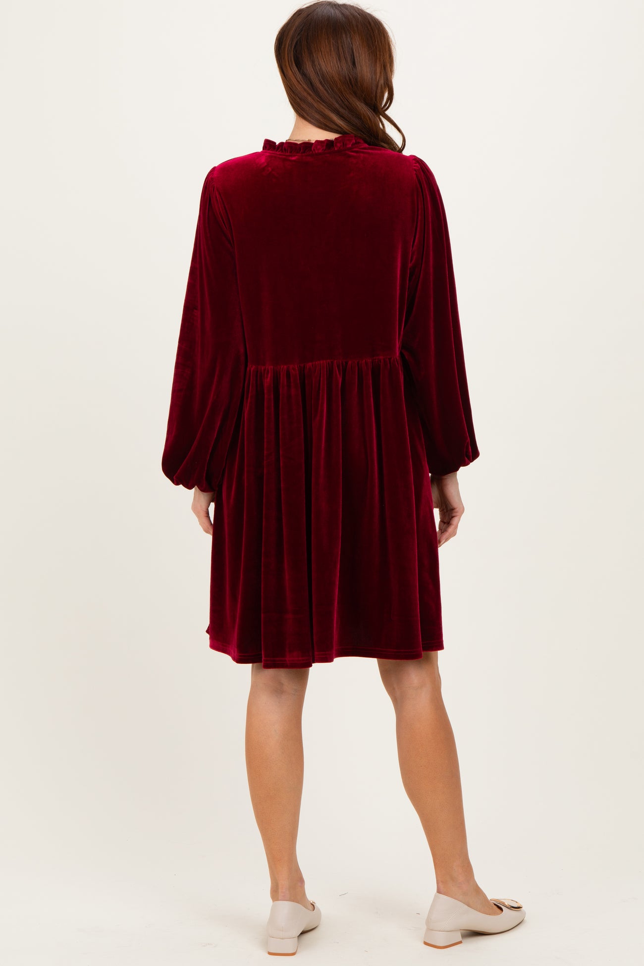 woman wearing a burgundy velvet long sleeve dress with a gathered waist and ruffled neckline, shown from the back