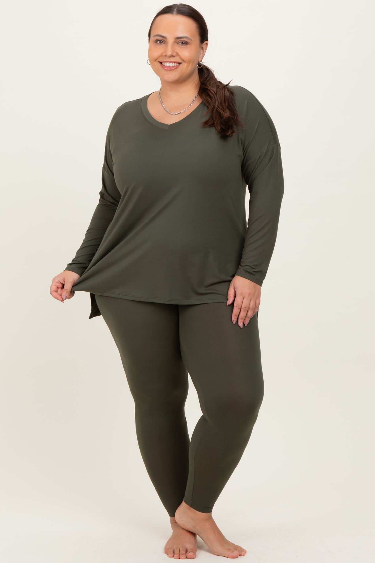 woman wearing a dark green long sleeve top and matching leggings, smiling while standing barefoot