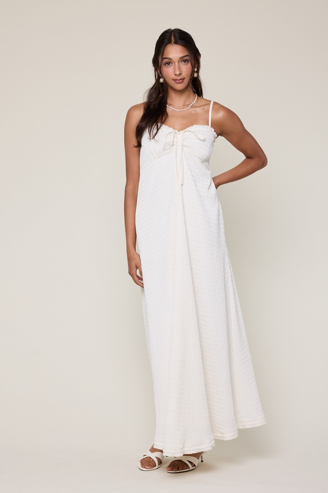 woman wearing a white maxi dress with bow detail and thin straps, standing casually with one hand on her hip