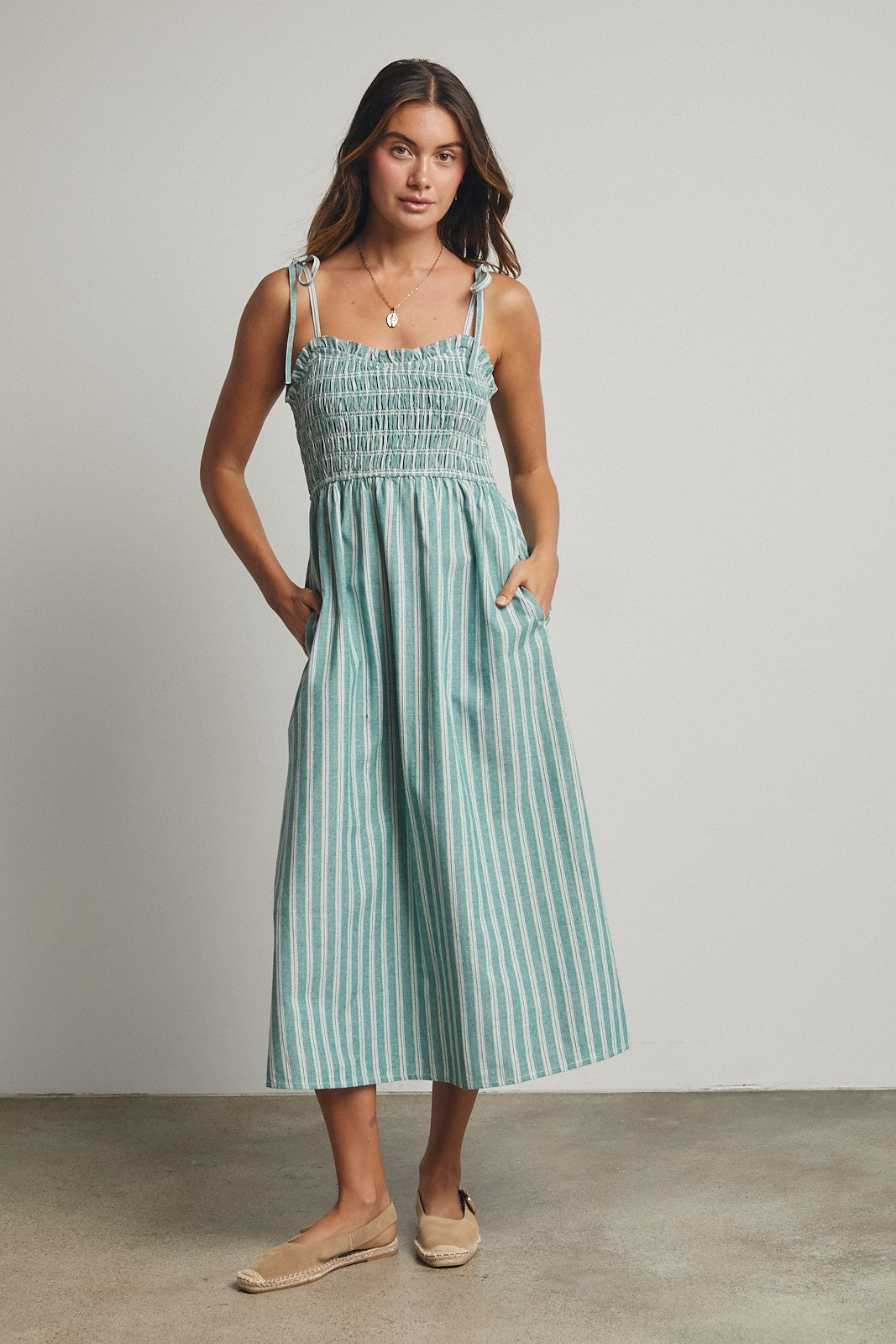 woman wearing a teal and white striped midi dress with smocked bodice and adjustable straps, standing with pockets