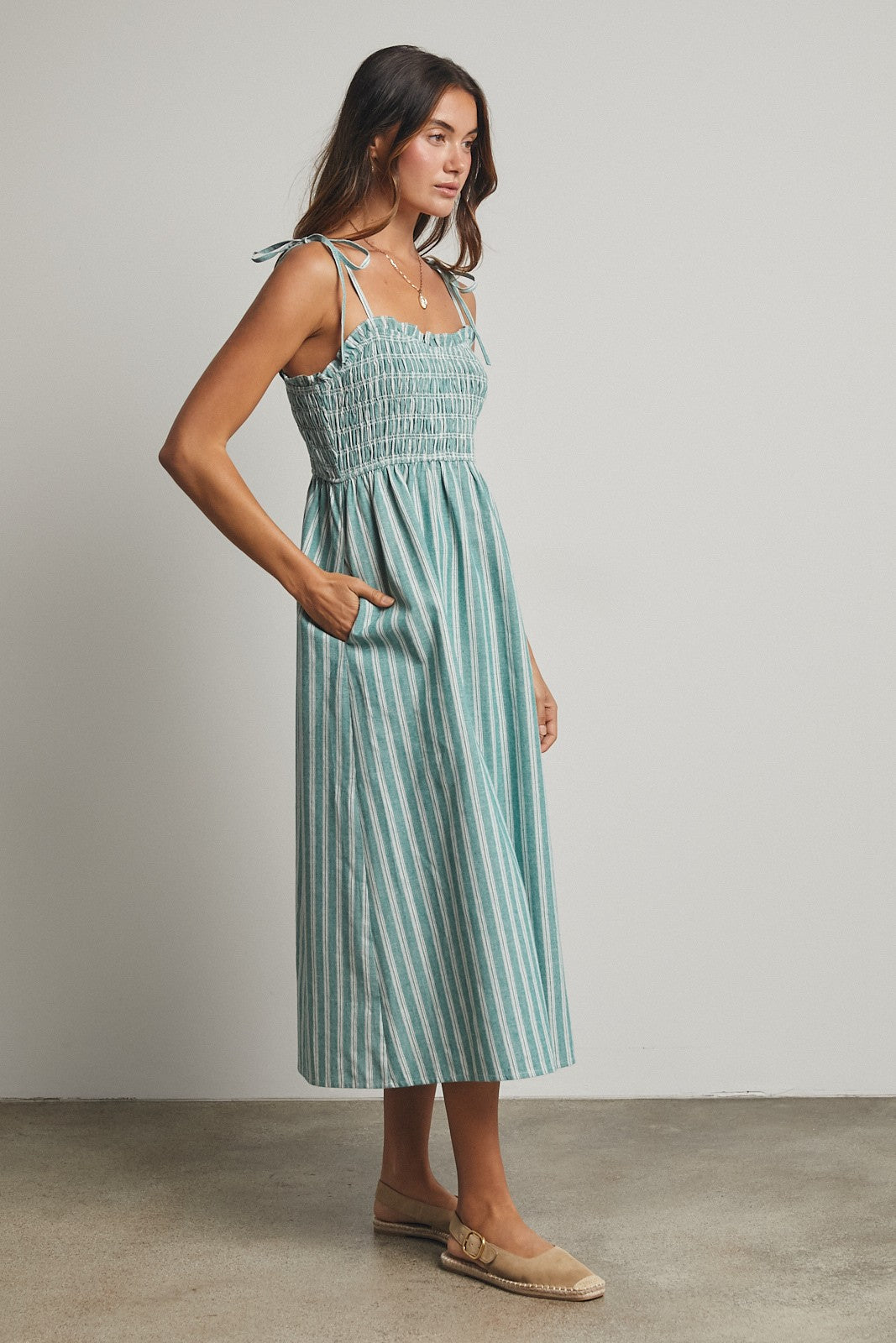 woman wearing a teal and white striped sundress with a smocked bodice and tie straps, featuring pockets, standing on a neutral background