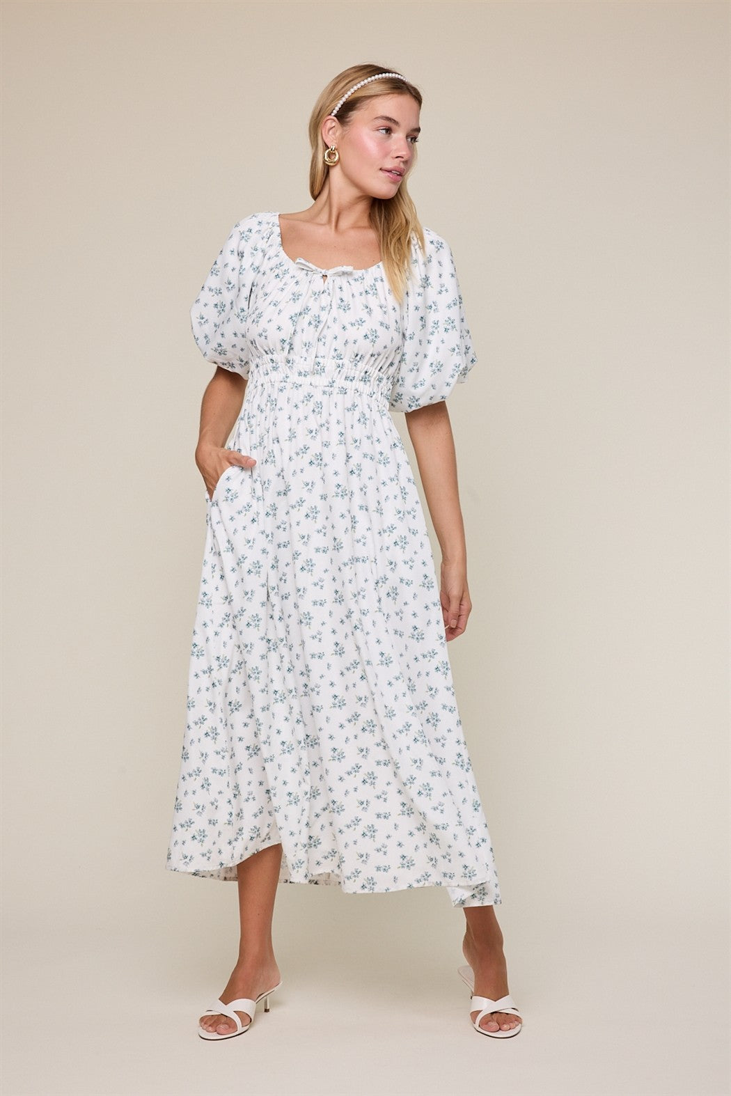 woman wearing a white floral maxi dress with puff sleeves and a cinched waist, posing with one hand in a pocket