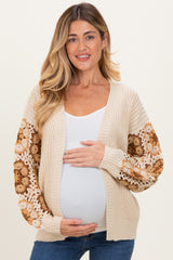 woman wearing a beige open front maternity cardigan with crochet floral sleeves and holding her baby bump