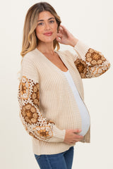 woman wearing a light beige maternity cardigan with floral knit detail on the sleeves and holding her baby bump