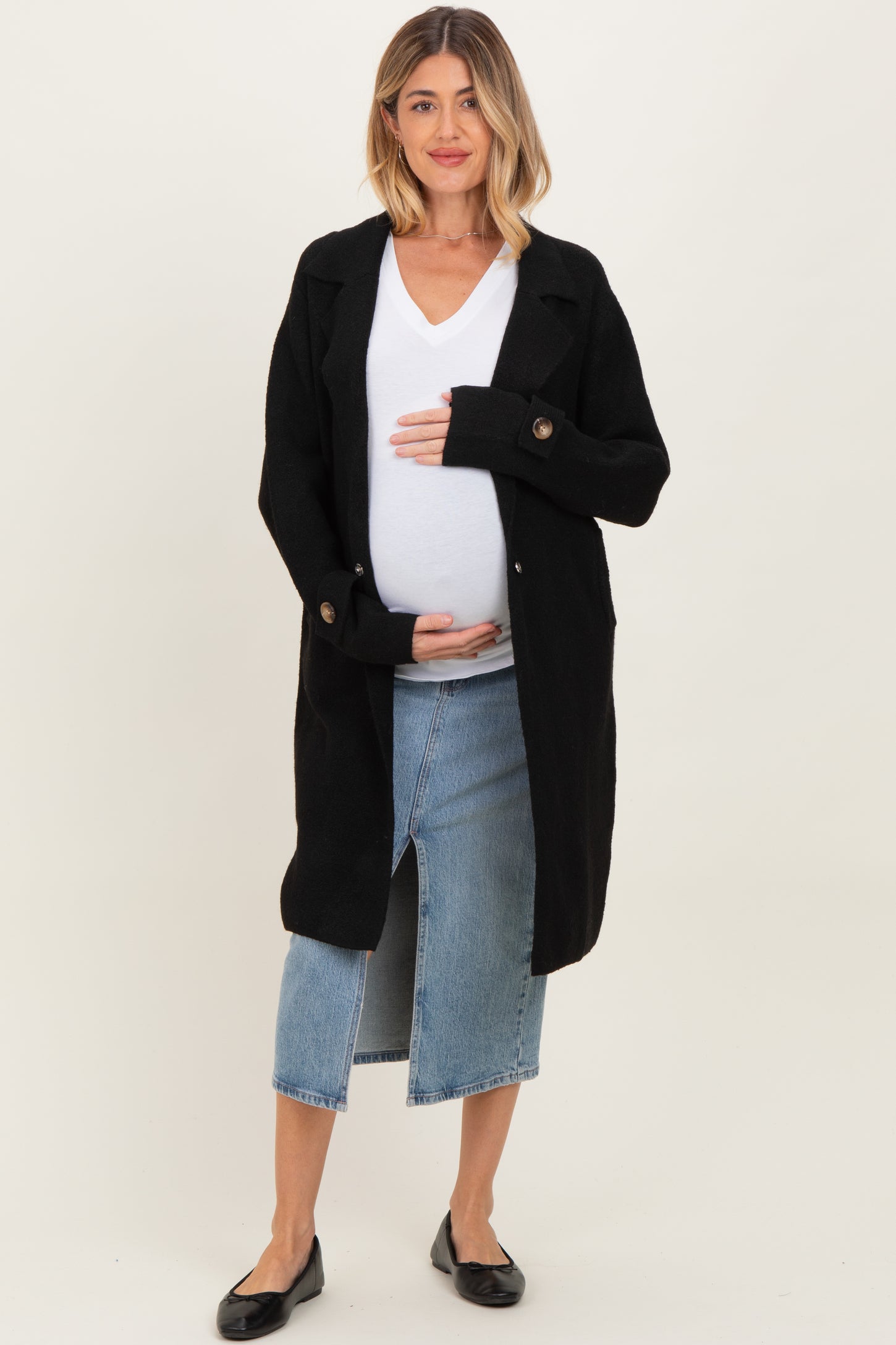 woman wearing a black long cardigan over a white v-neck shirt, holding her baby bump, paired with a denim skirt and flats