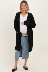 woman wearing a black long cardigan over a white v-neck shirt, holding her baby bump, paired with a denim skirt and flats