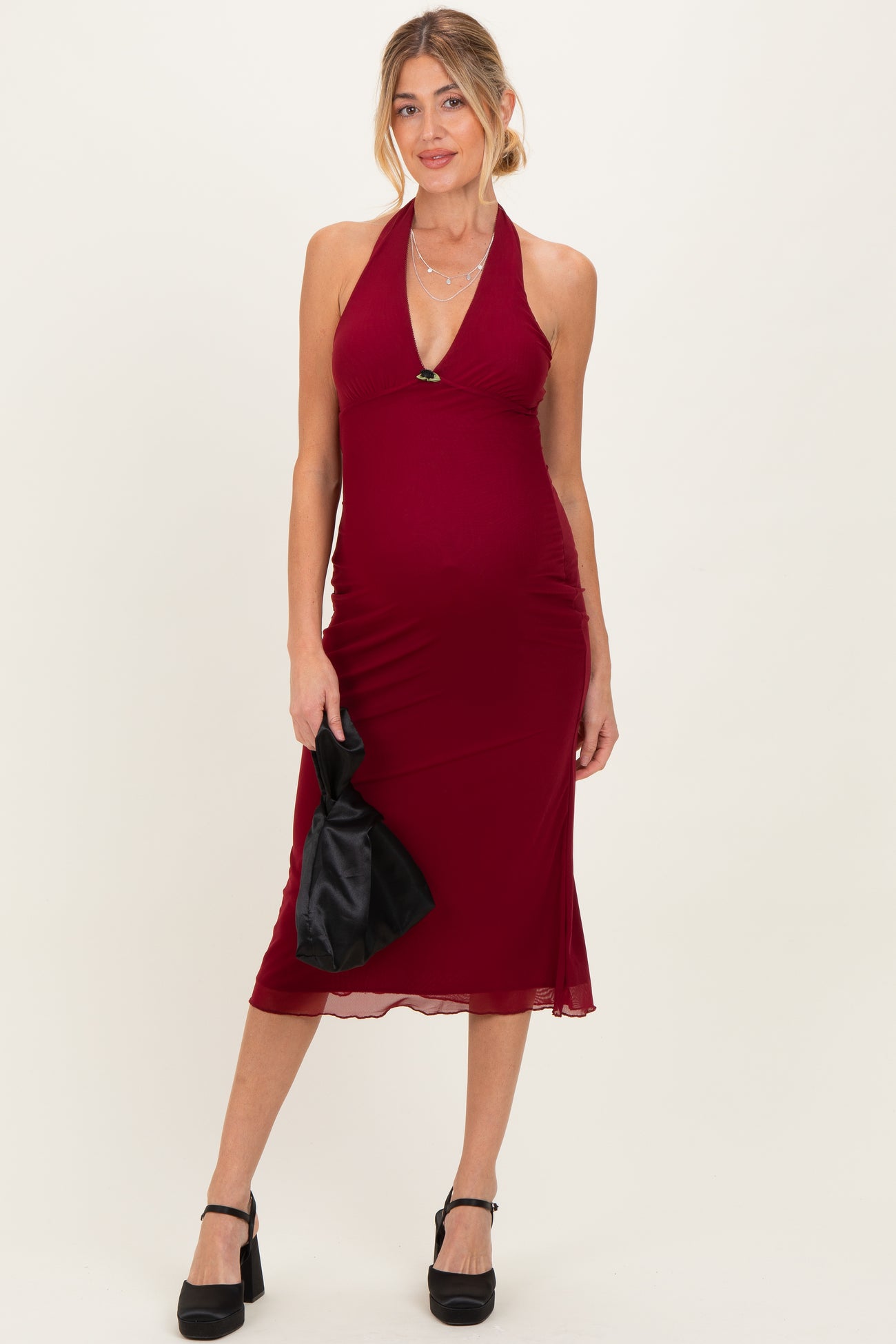 woman wearing a burgundy halter neck maternity dress showcasing her baby bump and holding a black clutch handbag