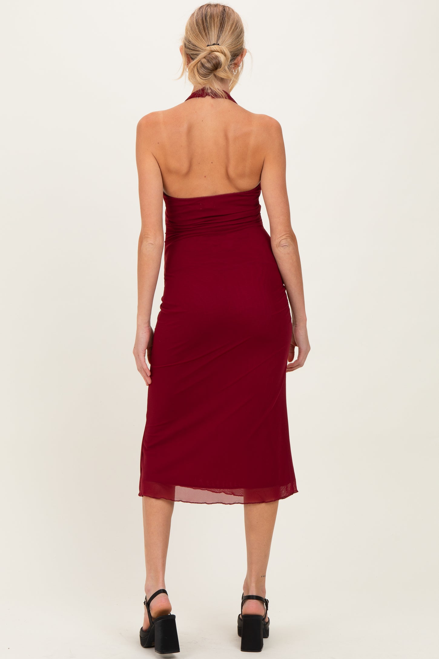 woman wearing a burgundy maternity halter neck dress with ruched details, showing a fitted silhouette from the back