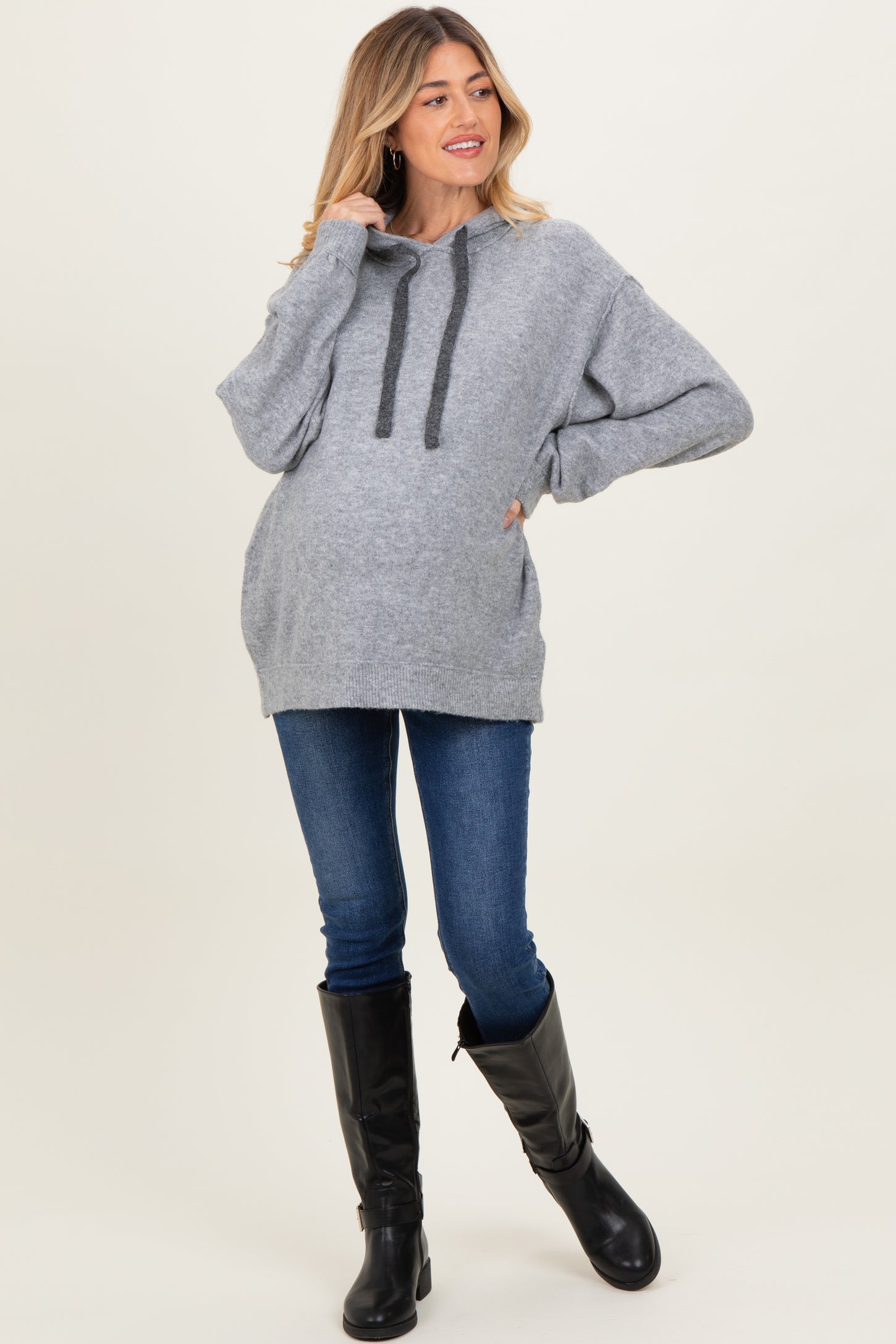 woman wearing a gray cozy maternity hoodie with a drawstring and pocket, paired with blue skinny jeans and black knee-high boots