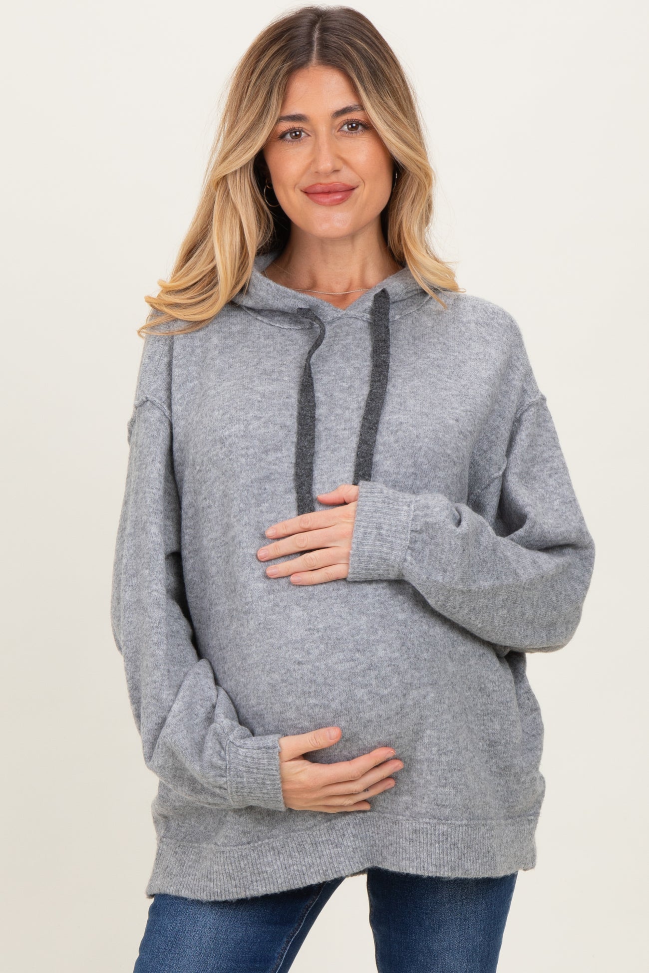 woman wearing a light grey oversized maternity hoodie, holding her baby bump with a smile