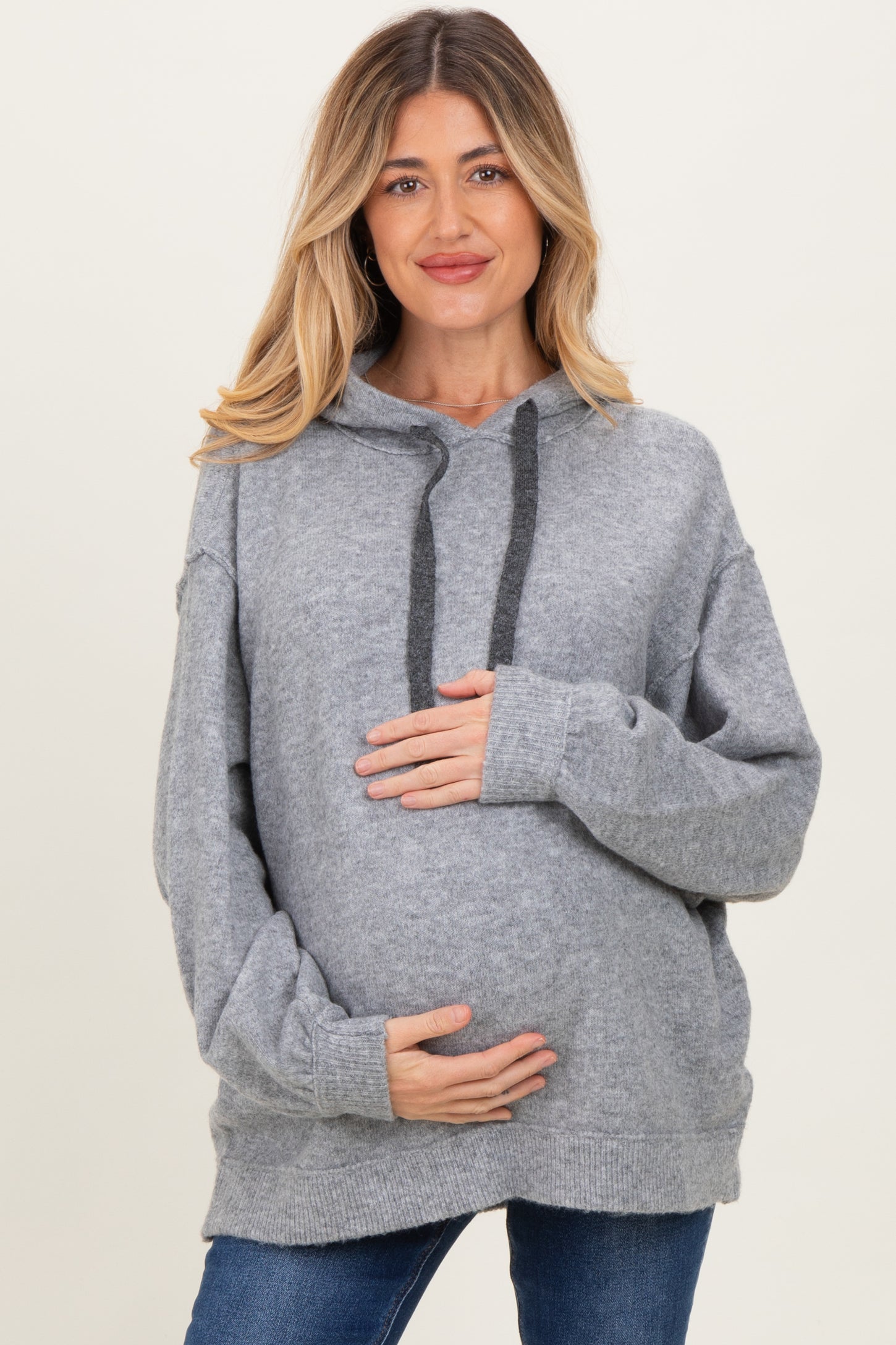 woman wearing a light grey oversized maternity hoodie, holding her baby bump with a smile