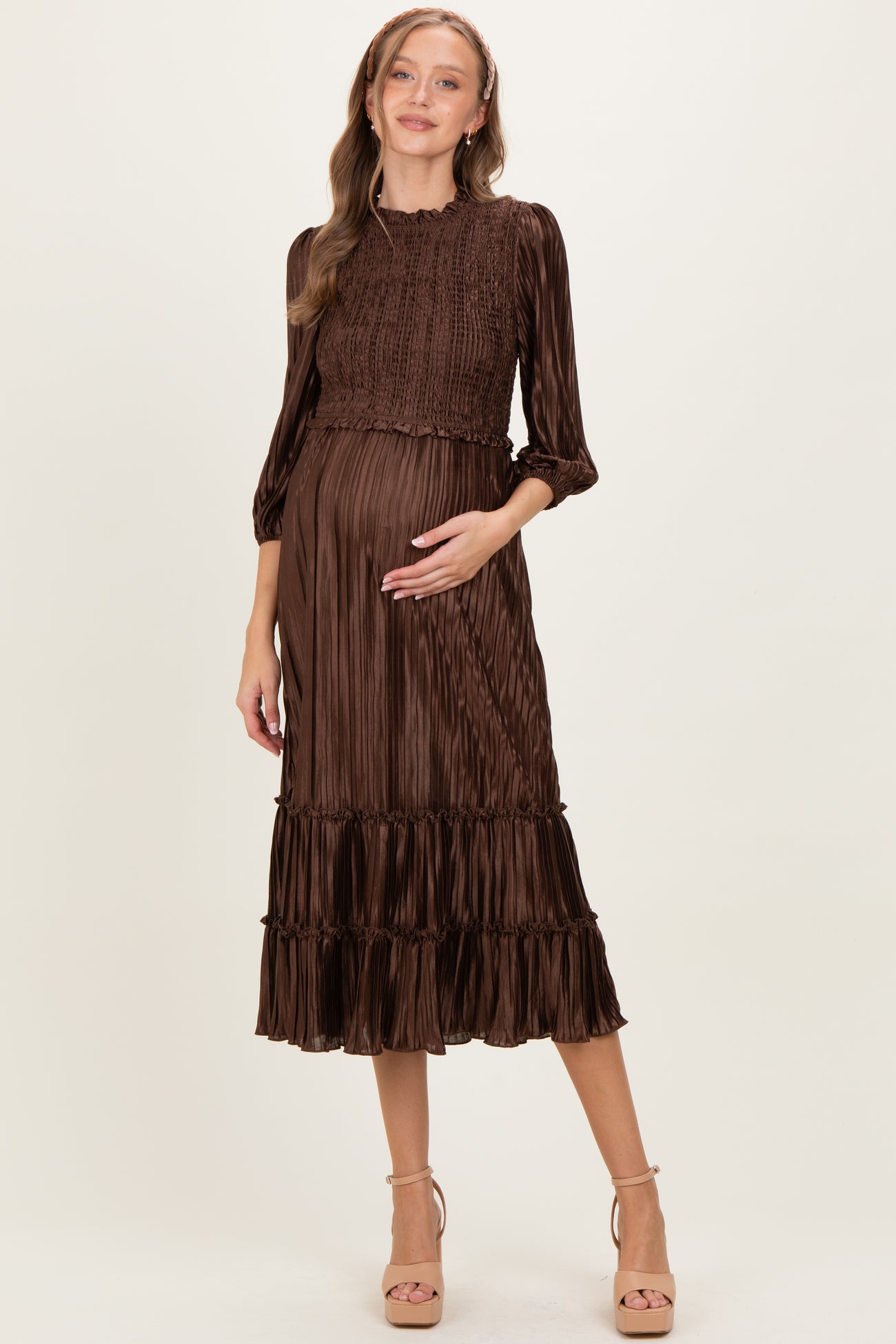woman wearing a brown pleated maternity dress with smocked bodice and ruffled hem, cradling her baby bump