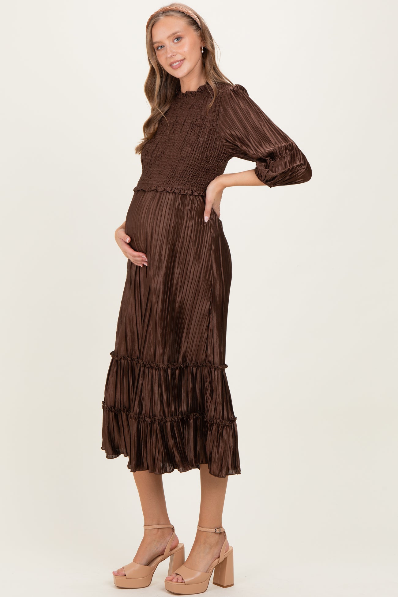 woman wearing a brown pleated maternity dress with smocked bodice and ruffled hem, holding her baby bump