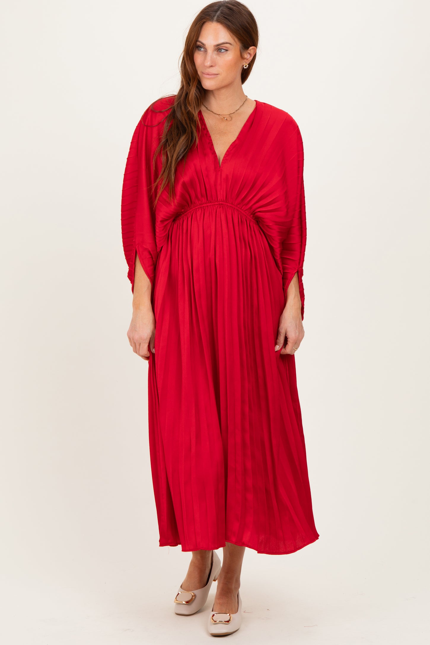 woman wearing a long red pleated dress with a deep v-neck and belted waist, standing against a neutral background