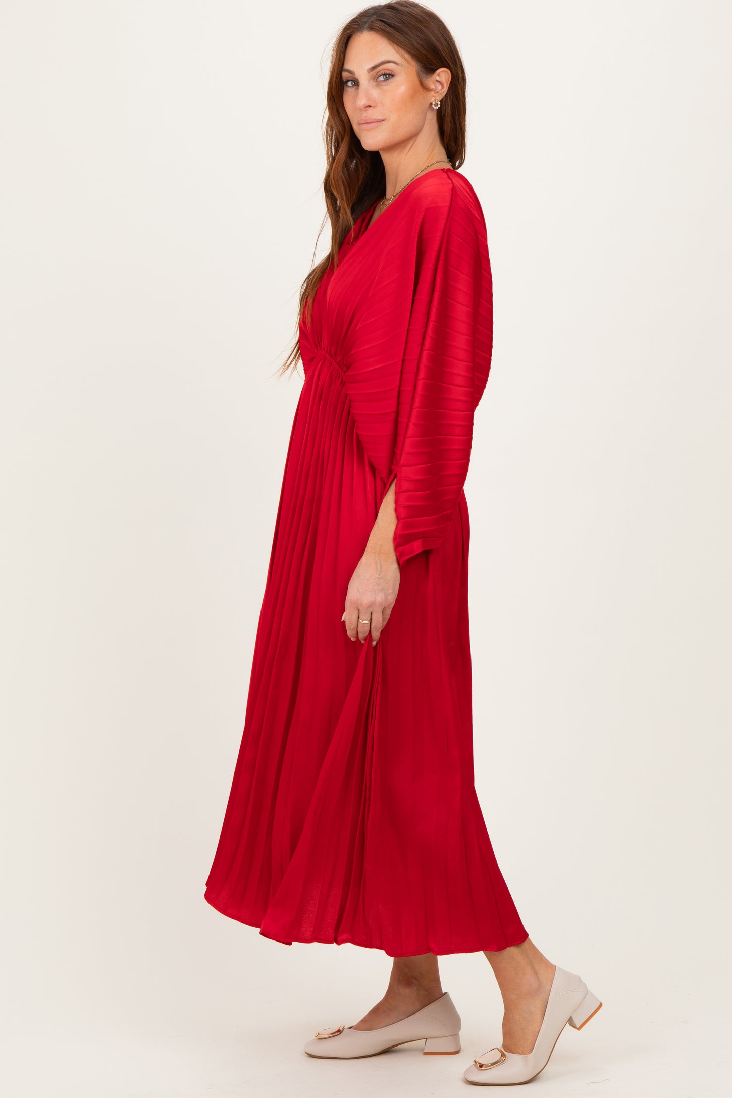 woman wearing a red pleated midi dress with long sleeves and a side knot detail, standing in profile