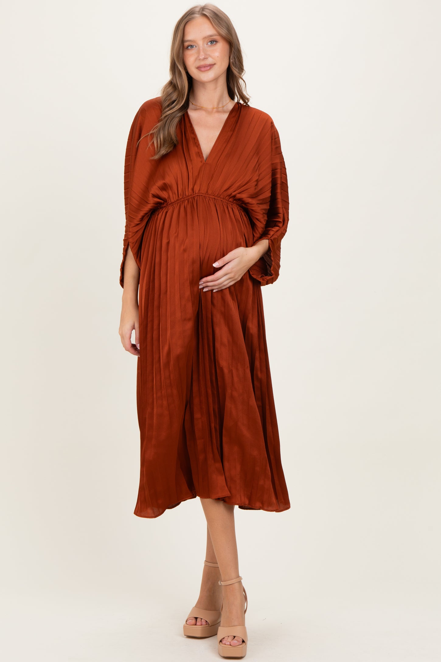 woman wearing a rust-colored maternity dress with a pleated skirt and v-neckline, holding her baby bump