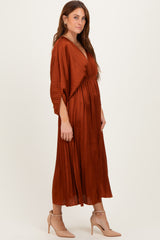 woman wearing a rust-colored pleated maxi dress with a deep v-neck and cinched waist, paired with beige heels