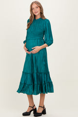 woman wearing a teal maternity dress with smocked bodice and tiered skirt, gently cradling her baby bump
