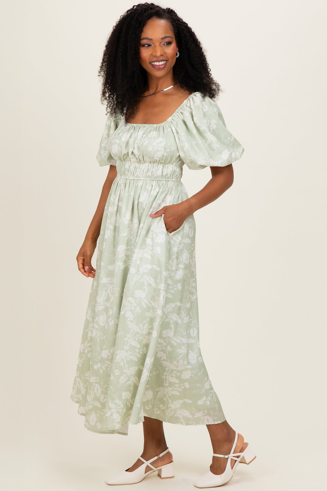 woman wearing a light green floral midi dress with puff sleeves and a cinched waist, standing with one hand in a pocket