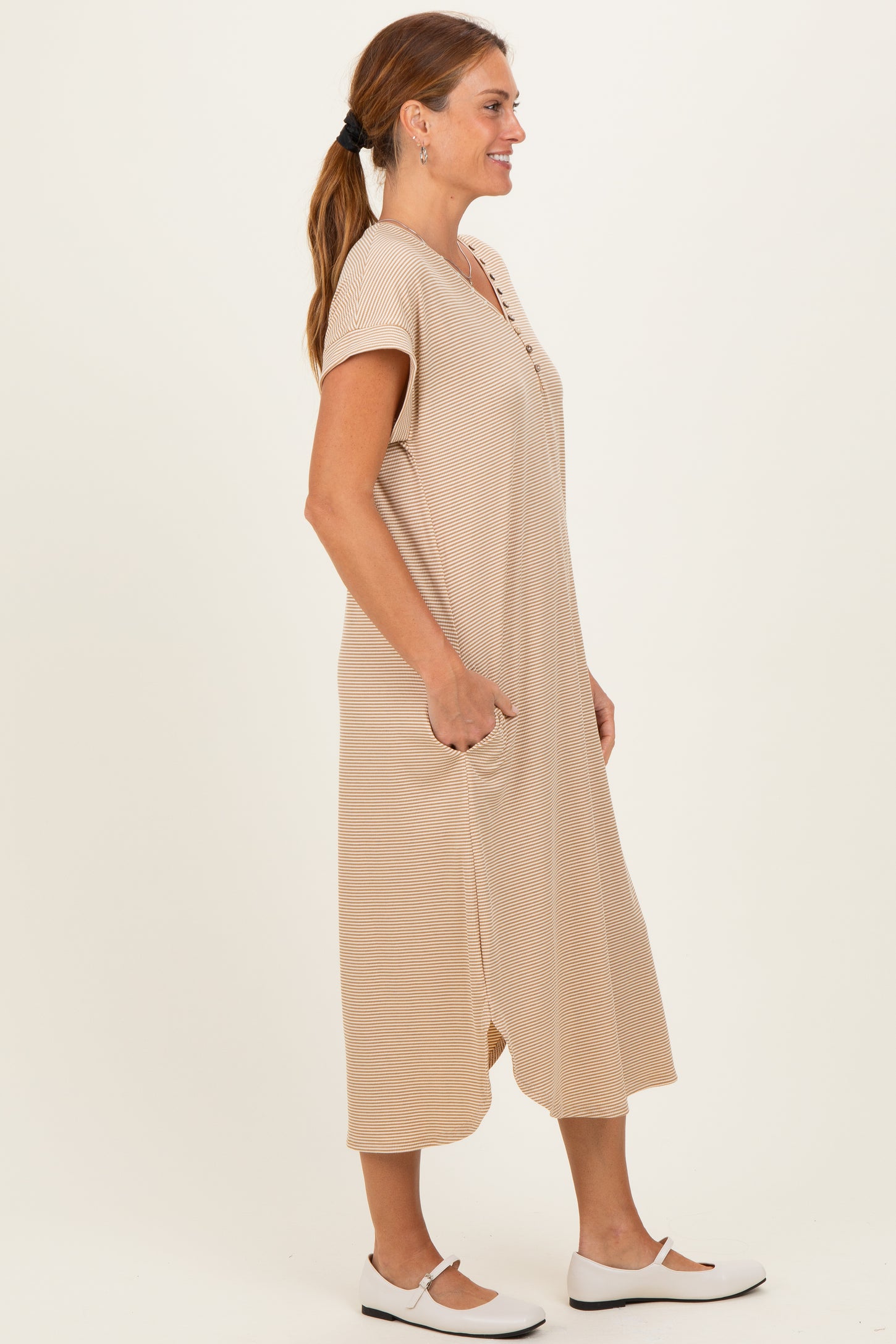 woman wearing a beige short sleeve midi dress with buttons and pockets, standing sideways against a neutral background