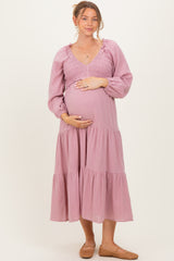 woman wearing a light pink long sleeve maternity dress with a smocked bodice and tiered skirt, holding her baby bump