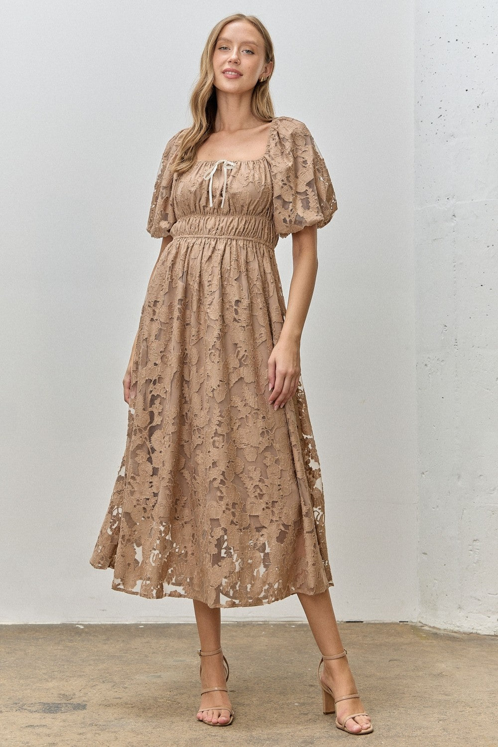 woman wearing a beige lace midi dress with puff sleeves and a cinched waist, standing on a concrete floor