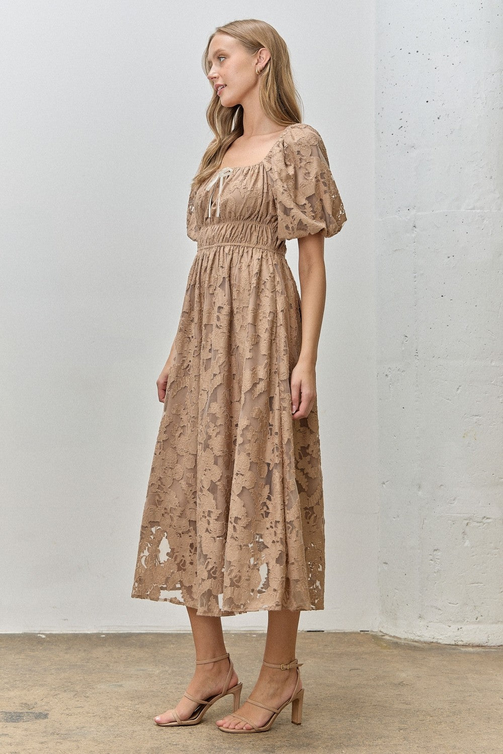 woman wearing a brown floral lace midi dress with puff sleeves and a cinched waist, standing against a light background
