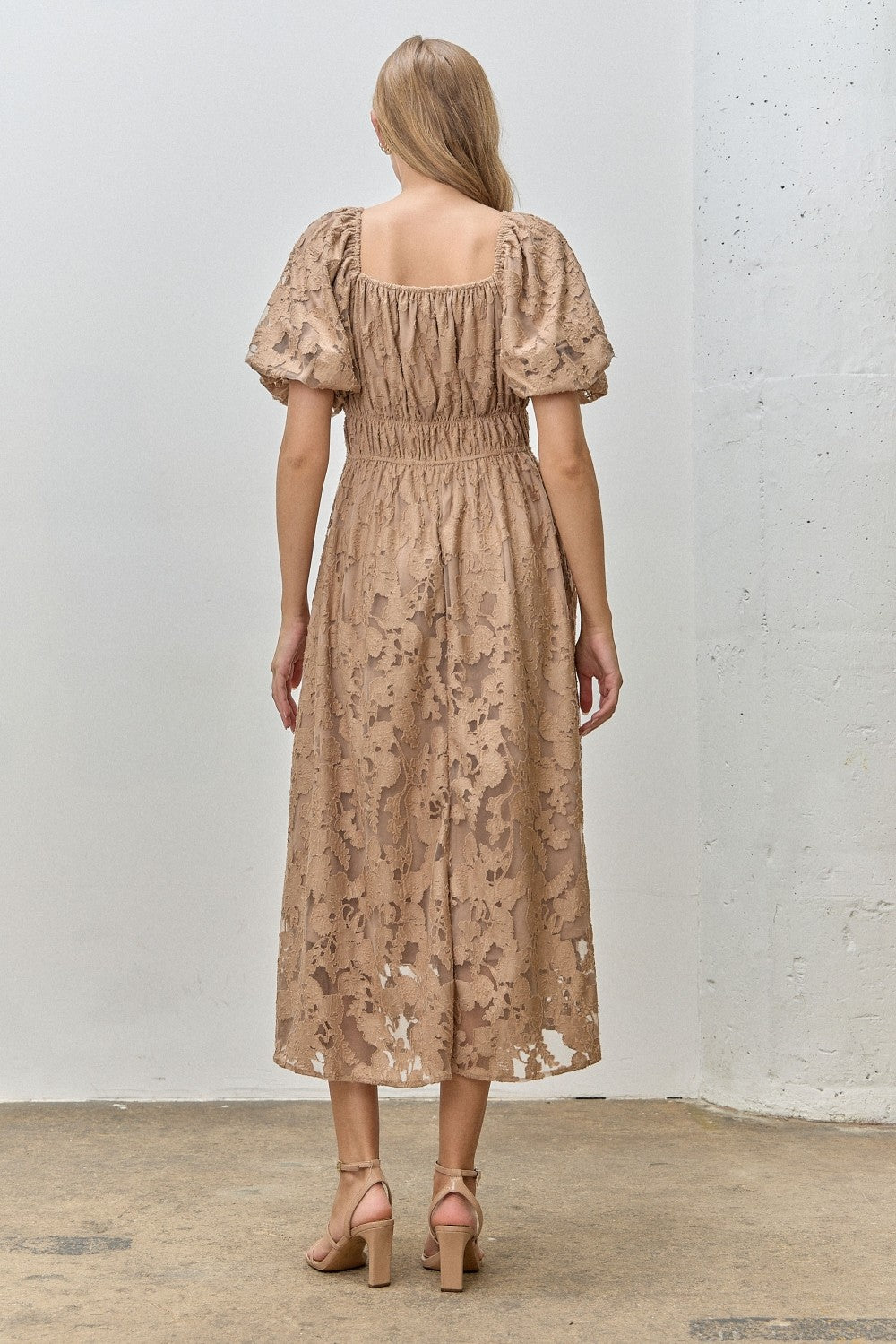 woman wearing a beige puff sleeve midi dress with a fitted waist and floral lace detailing, shown from the back