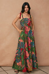 woman wearing a colorful floral maxi dress with tied straps and pockets, standing against a textured backdrop