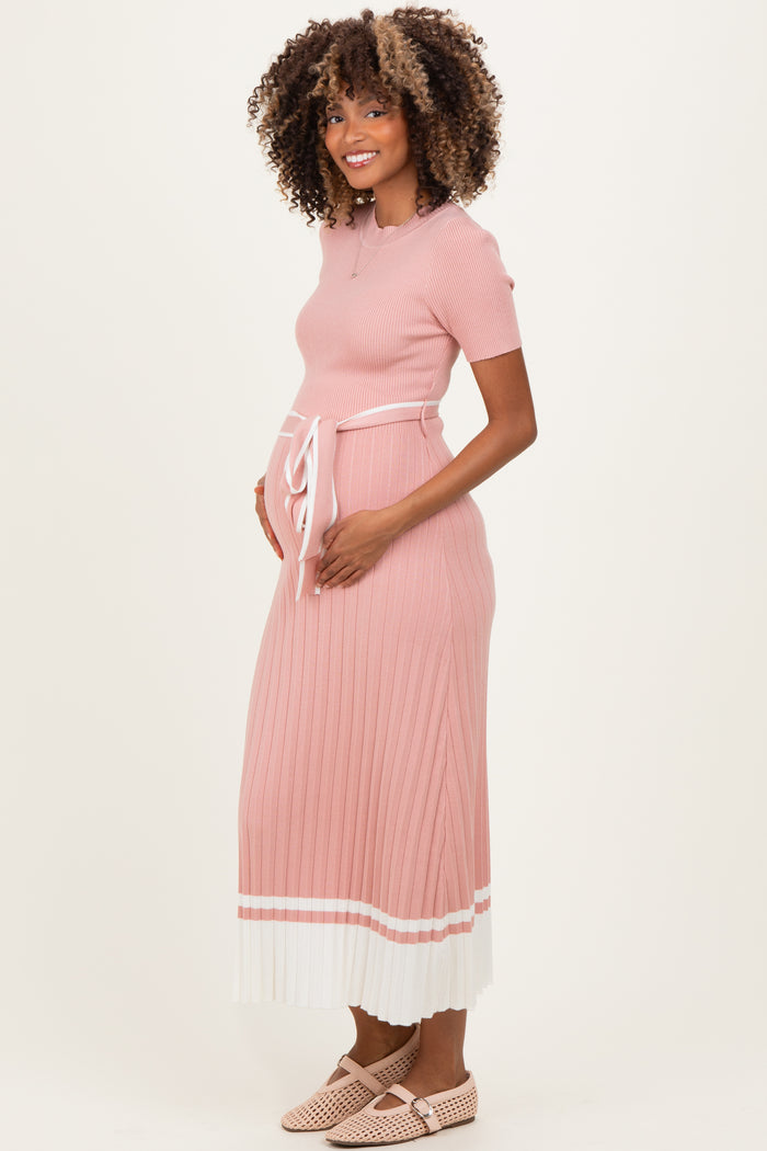 woman wearing a pink ribbed maternity dress with short sleeves, a tie waist, and a pleated skirt, holding her baby bump