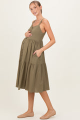 woman wearing an olive green maternity dress with adjustable straps, a tiered skirt, and pockets, showcasing her baby bump